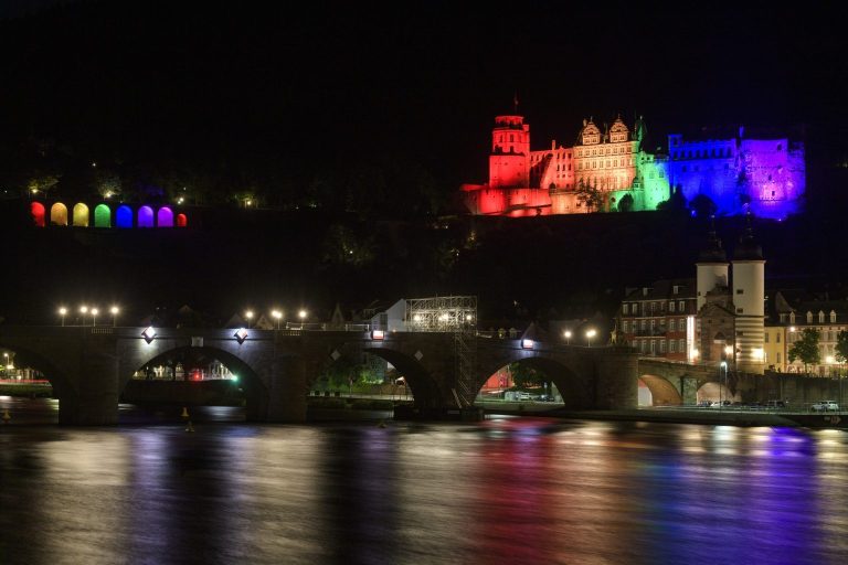 Heidelberg in bunten Lichtern bei Nacht, reflektiert im Wasser des Flusses.
