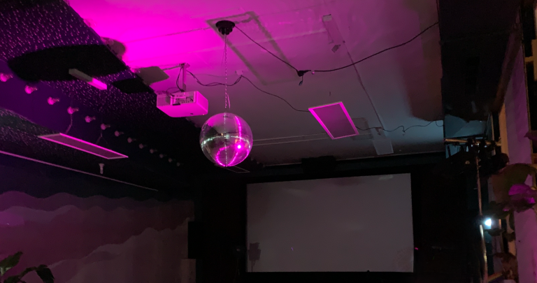 Queer Space Heidelberg Eine Discokugel hängt von der Decke, umgeben von pinkem Licht und einer Leinwand.